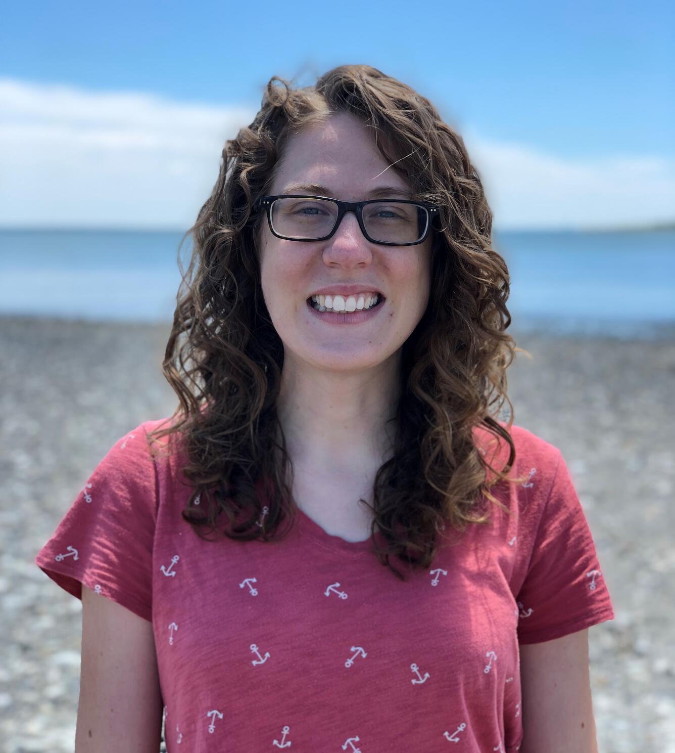 Photo of Jennifer Meineke, wearing a coral-colored shirt. She is smiling and standing in front of a blue sky. 