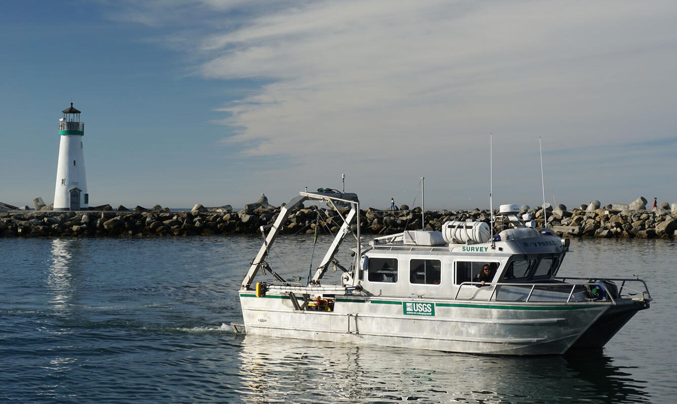 A small boat motors in a calm waterway with a rocky jetty behind, where a lighthouse sits in the distance.