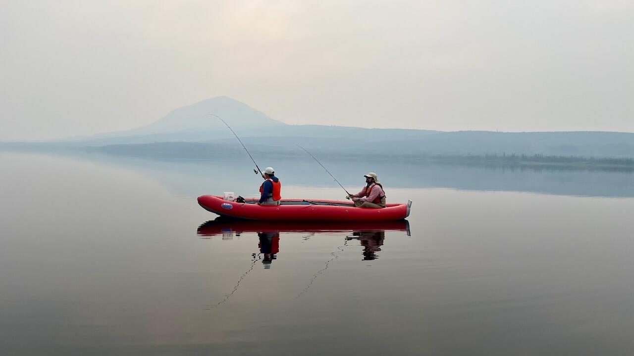 Female and male sitting in red inflatable skiff on lake with fishing poles. Both wearing hats and orange PFDs.