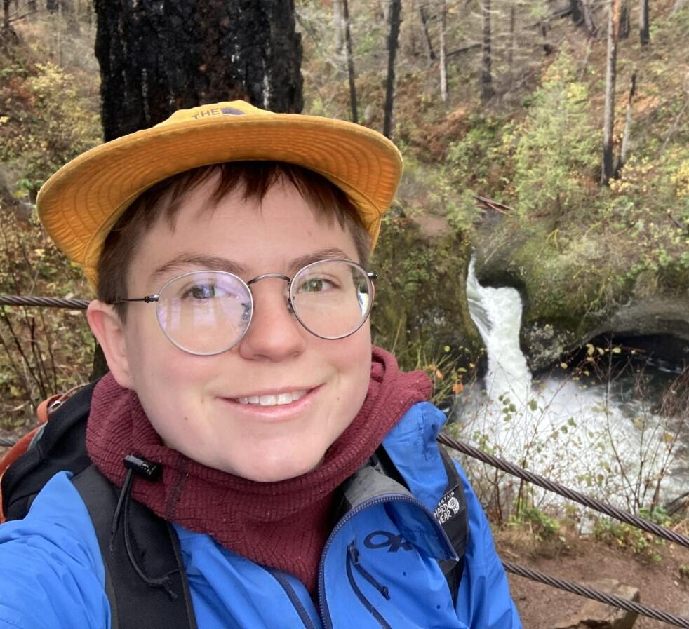 A photo of USGS Hydrologist. Julia smiles wearing in front of a partially burned forest area hillside with stream.