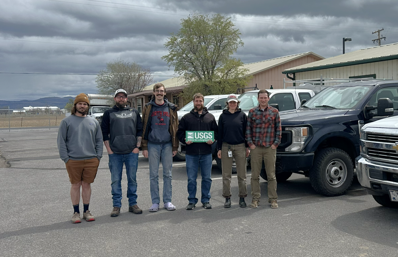 Six people stand in a row in a parking lot with trucks outside single story rectangular buildings on a cloudy day