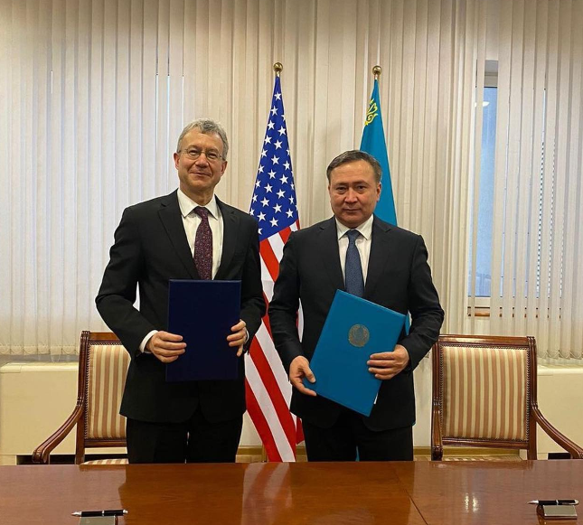 Photograph of two officials with flags, pens, and a document at a table in a room with curtains