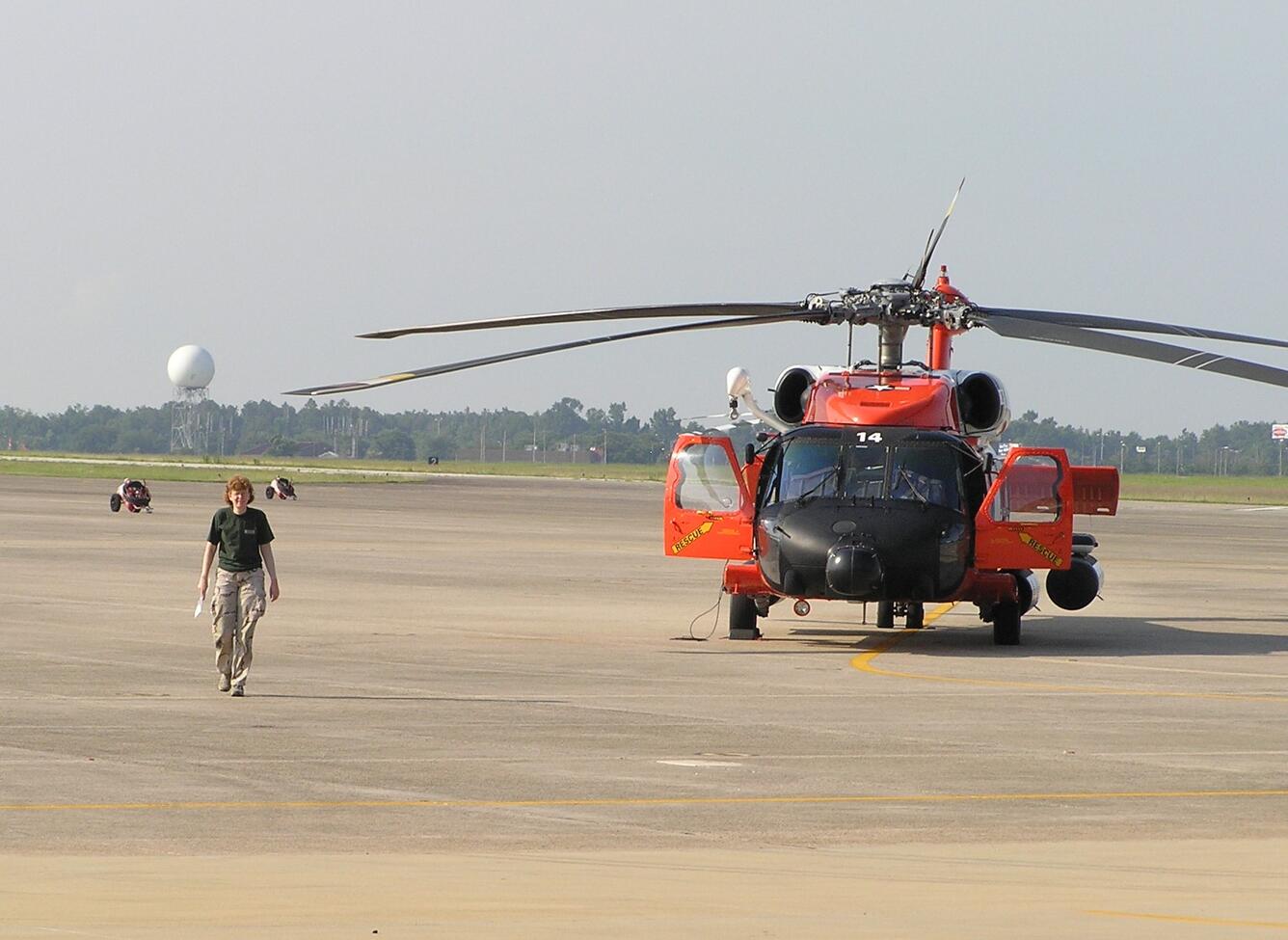 A woman walks towards the camera and away from a large black and orange helicopter on a large landing pad.