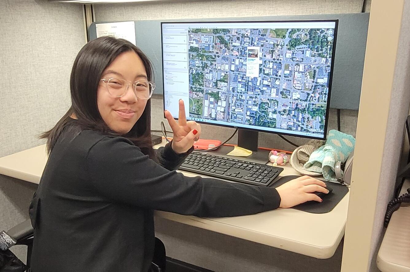 A smiling woman with glasses and wearing a black shirt flashes a peace sign while working on a computer.