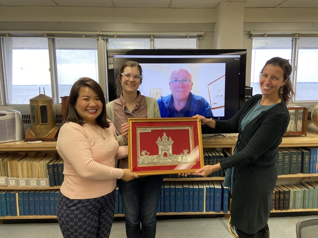 Three smiling women are holding a framed photo, with one man on a computer screen.