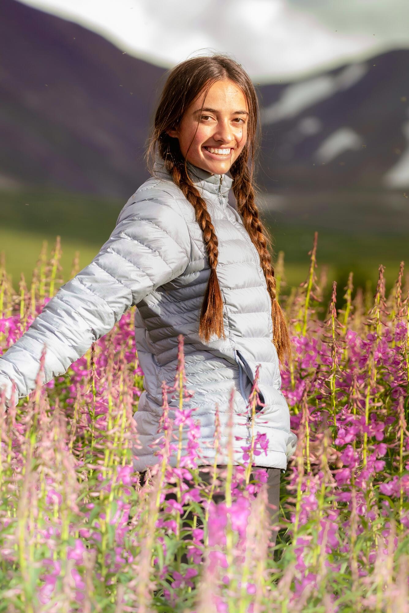 Person with long hair in two braids standing among a field of wildflowers with mountain in background