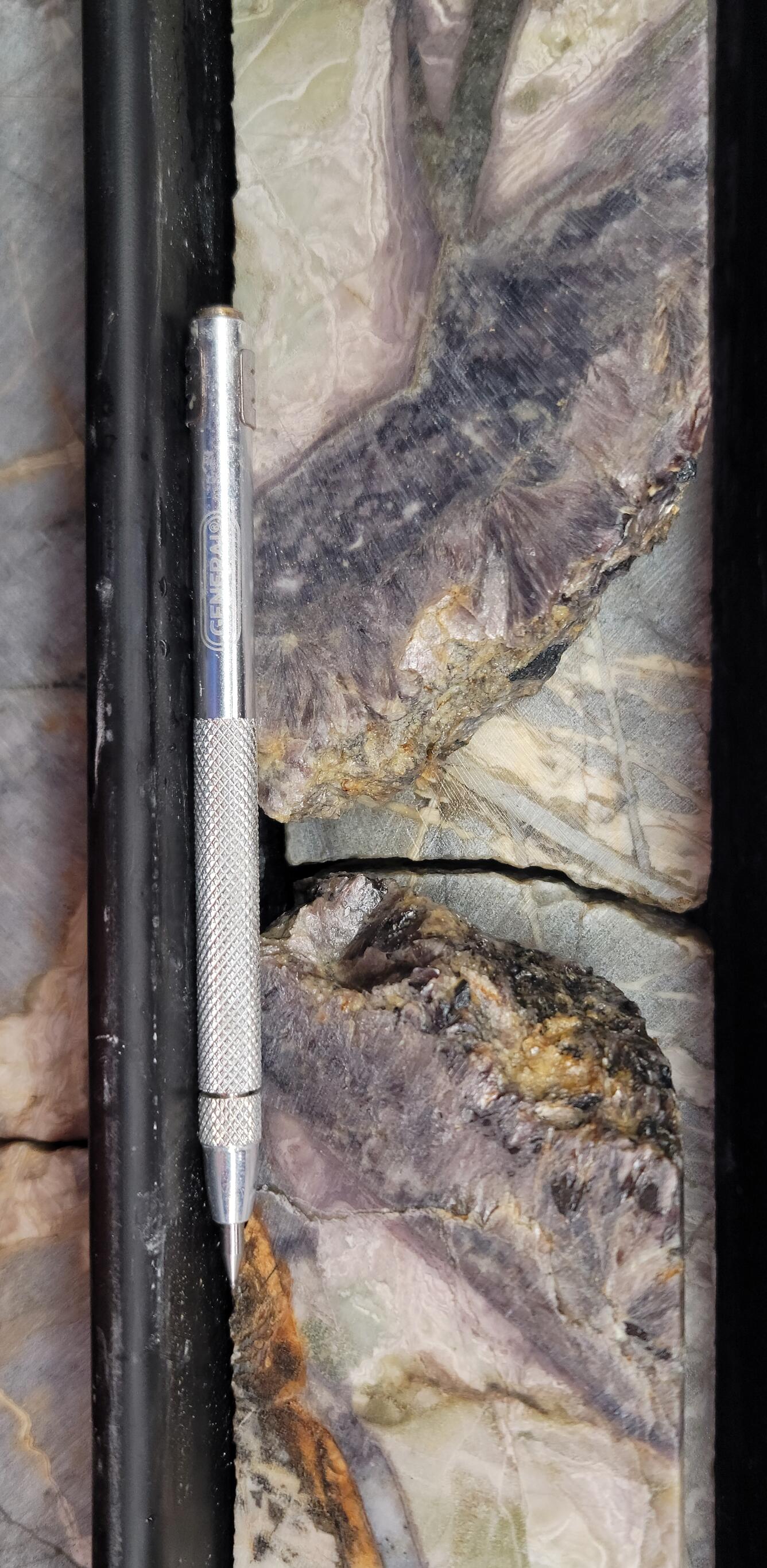 Gray with bits of white cylinder-shaped vein intersected in drill core. Silver pencil next to core for size.