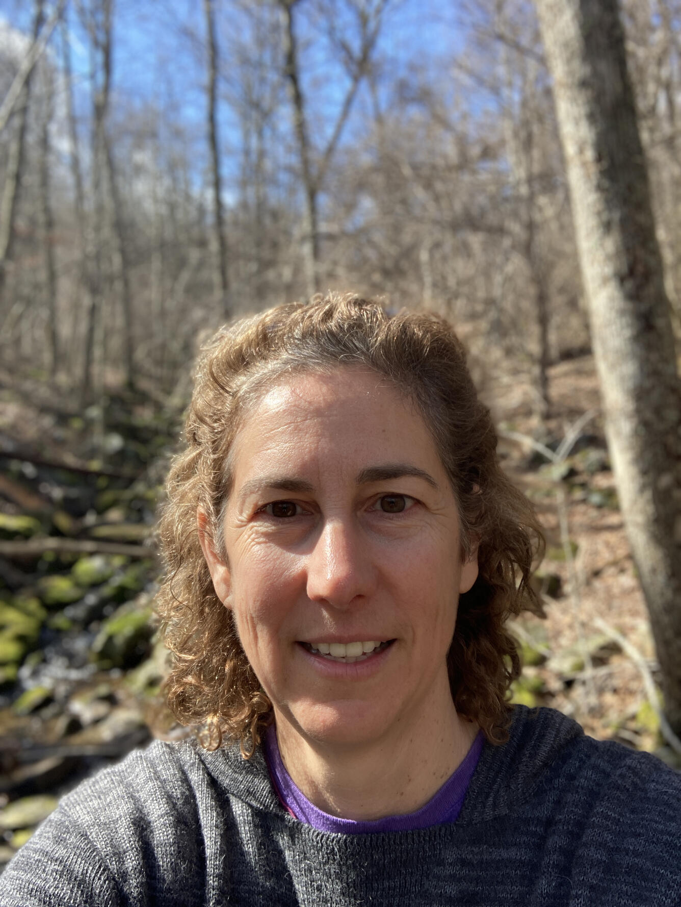woman wearing a gray sweatshirt standing in the woods on a sunny day