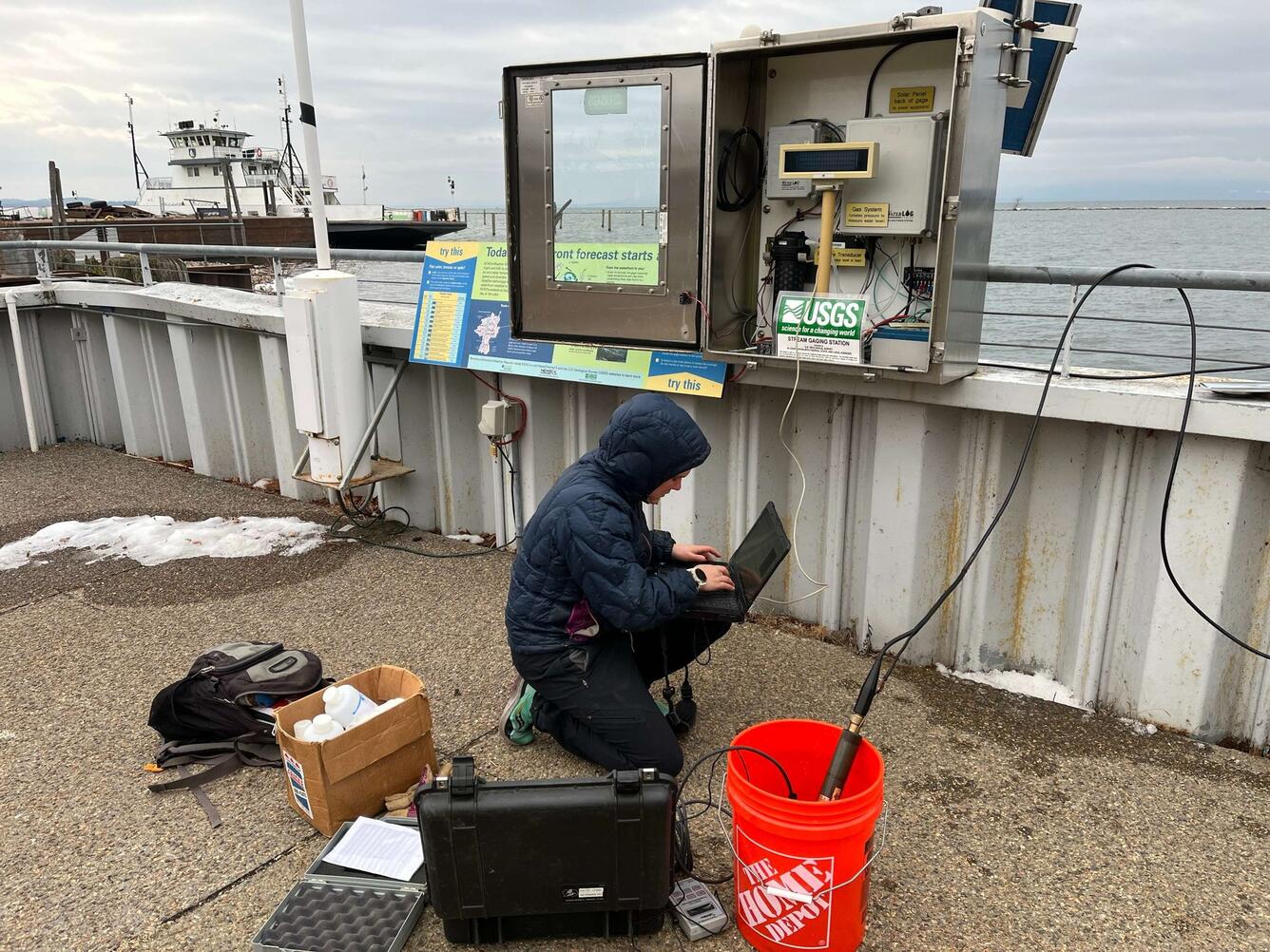 Hydrologic technician squatting down with a laptop next to a streamgage connected to a waterfront railing.