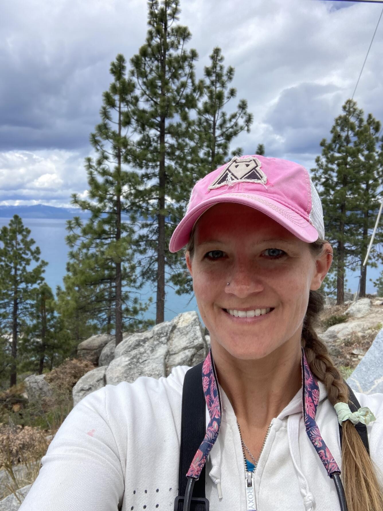 Headshot of a woman, wearing a white zipped jacket and pink baseball cap, with tall pine trees and a lake in the background