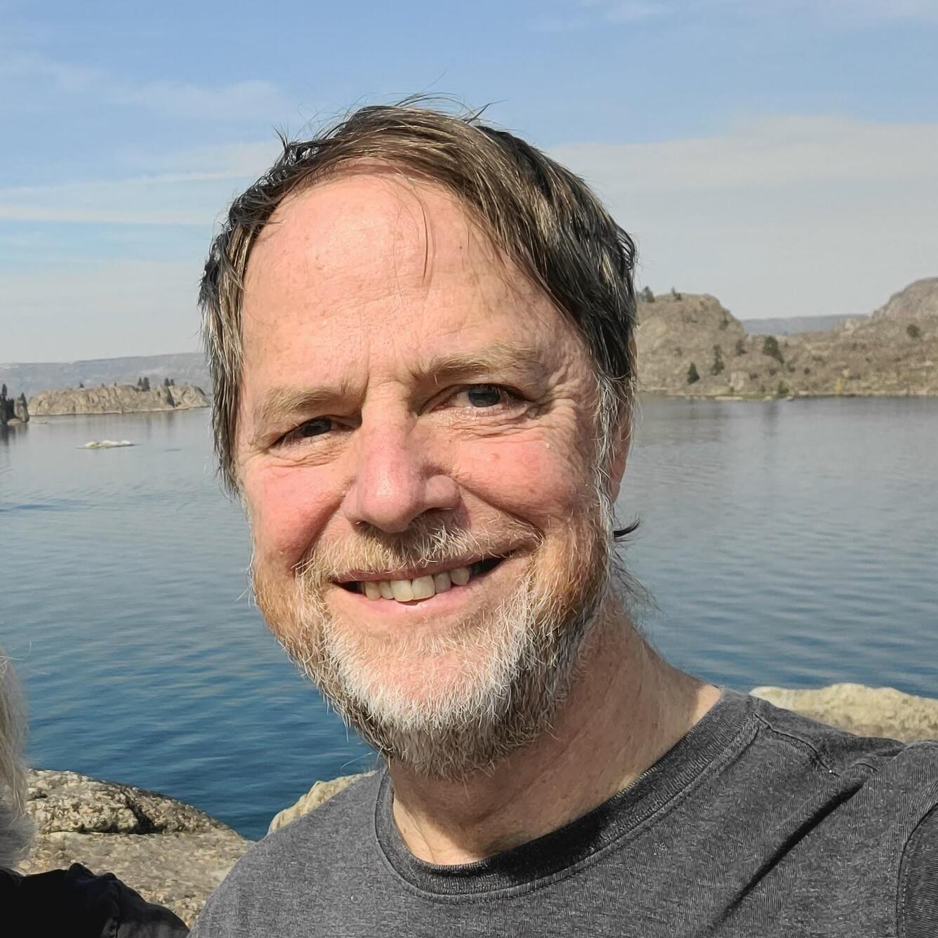 Man with brown hair and white beard smiling with a lake in the background.