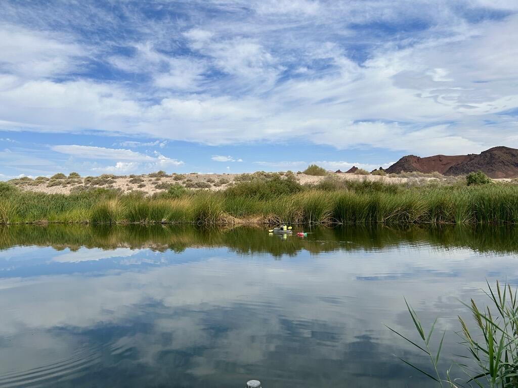 Person in yellow kayaking near far bank on Las Vegas Wash, towing stream gaging equipment under partly cloudy sky.
