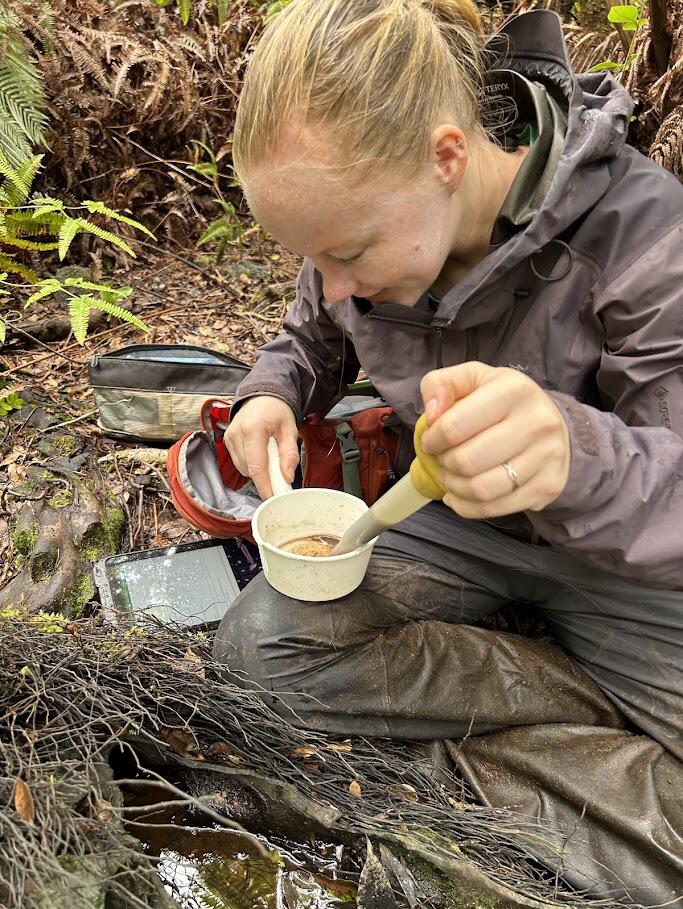 Person sitting on ground in rain gear holding a sample in a plastic cup and a baster.