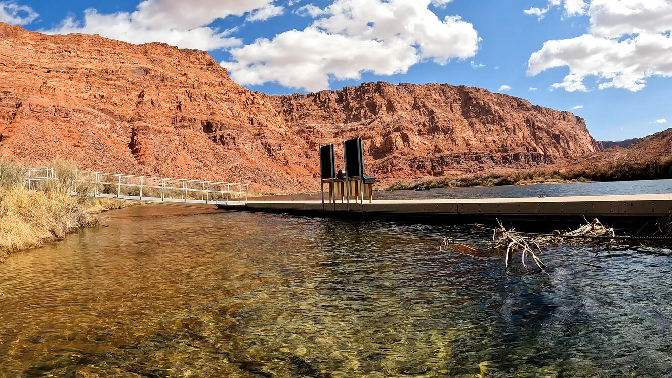 A long dock stretches out over a calm river, with tall red canyons in the background.