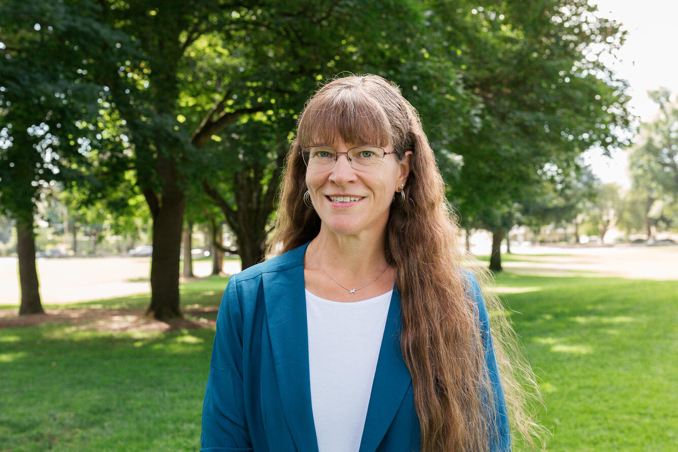 A woman with long brown hair and glasses is wearing a white shirt with a light blue jacket as she smiles for her picture in a park.