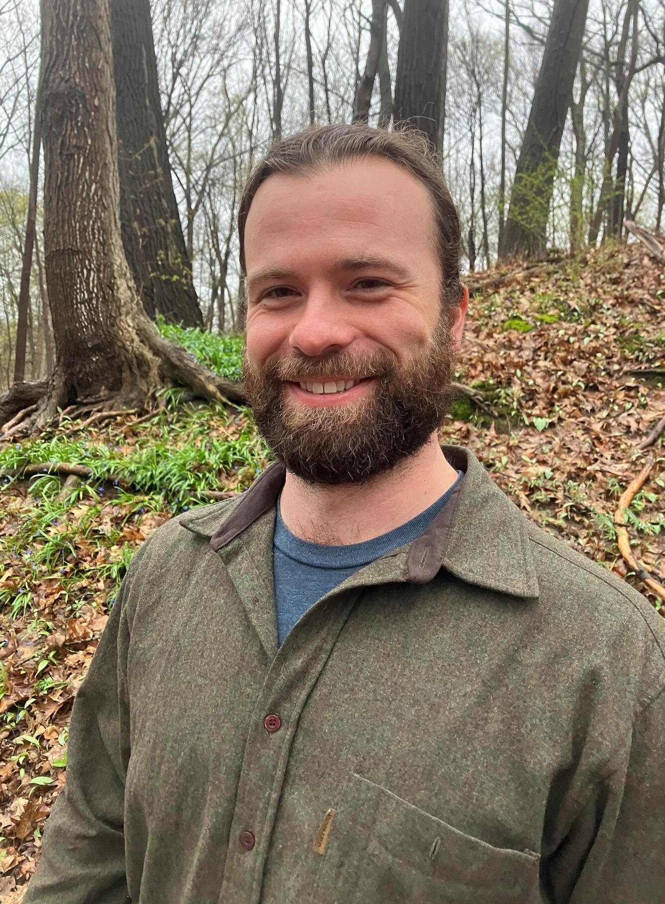 A man with a beard and a green shirt is smiling and standing in a forest.