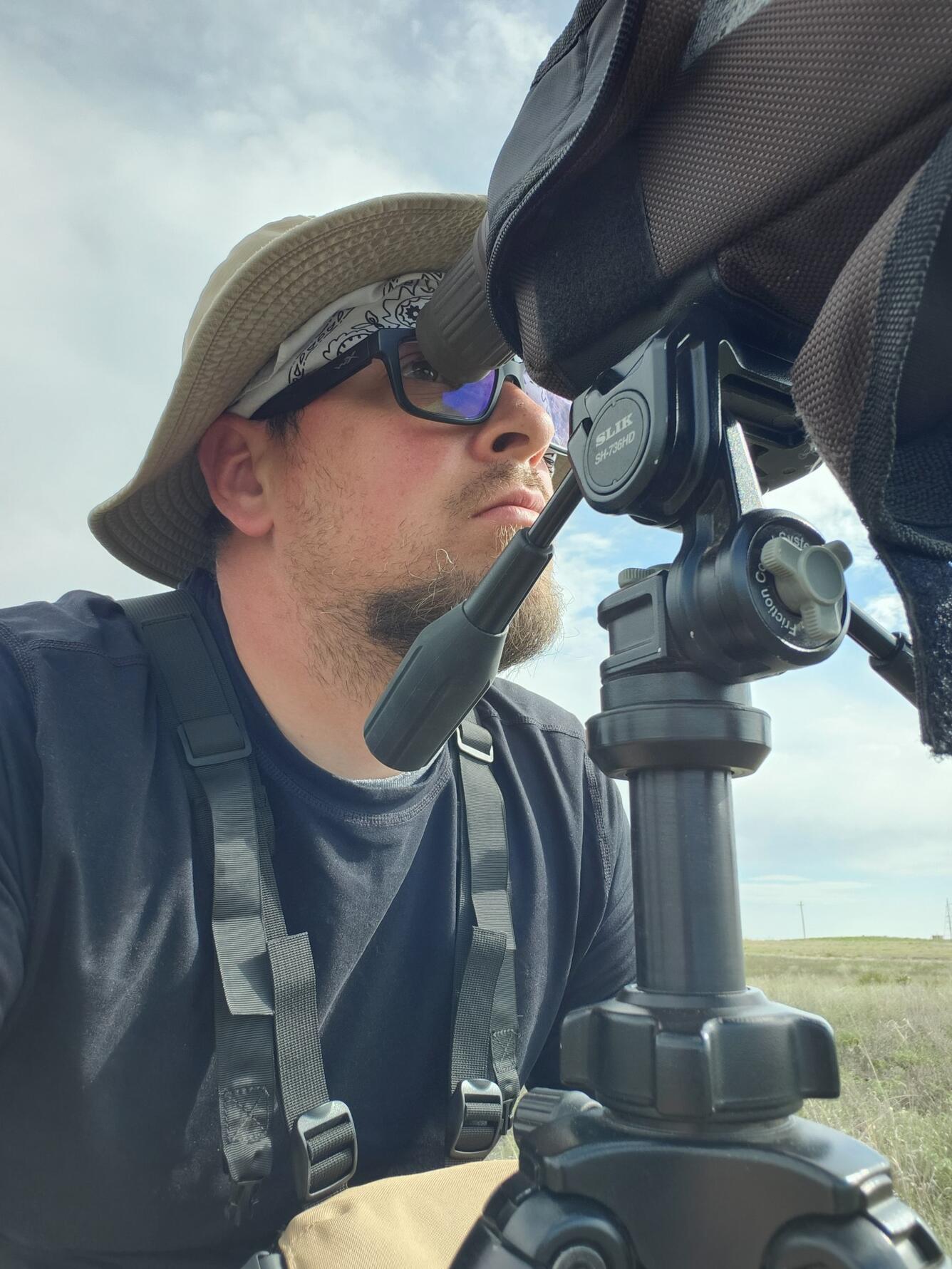 man wearing sunglasses, blue shirt, tan pants looking through a tripod on a partly sunny day