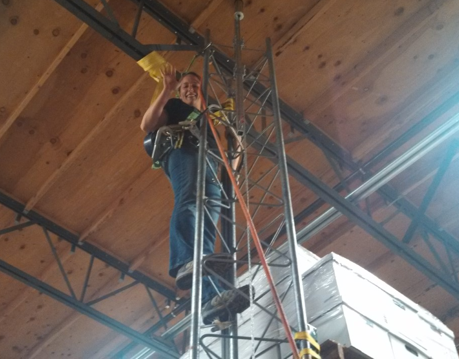 Safety officer Lori demonstrating fall safety waves down from a tower that she has safely climbed with a harness and gear.