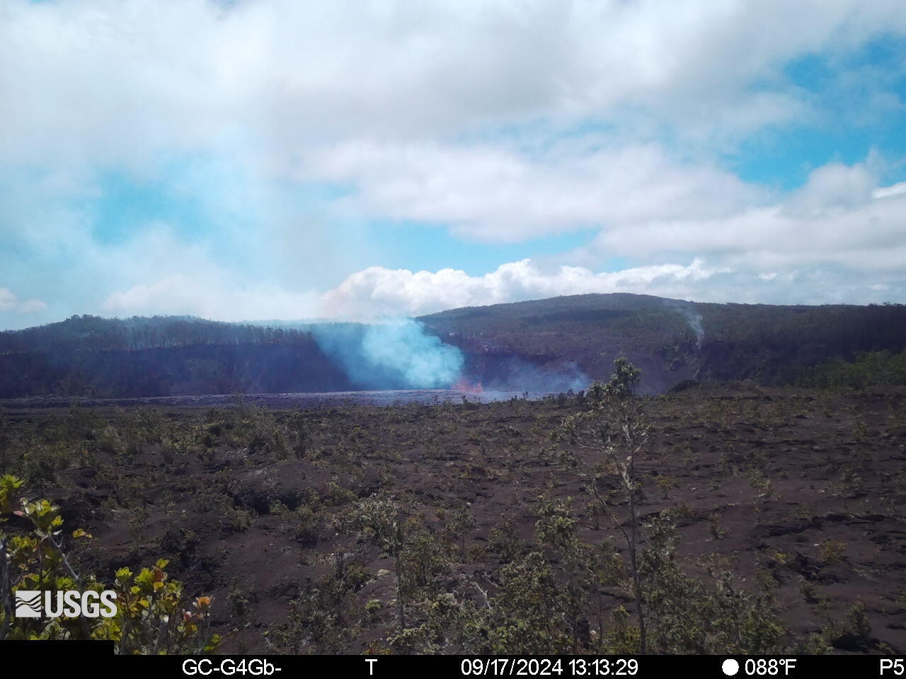 Color webcam view showing glowing lava and a volcanic plume in the background