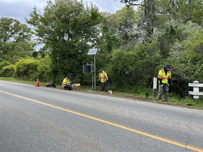 Three USGS staff in neon yellow safety vests installing a streamgage on the side of the road.