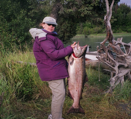 woman wearing a purple jacket, sunglasses, and gray hat holding a fish near a stream and trees on a sunny day