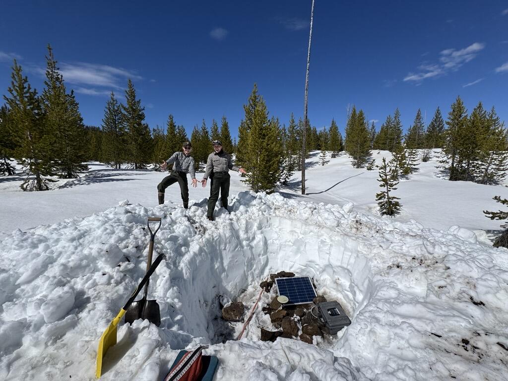 Two woman point out a pit in snowy landscape with equipment in the pit. Sparse trees and blue sky in background.