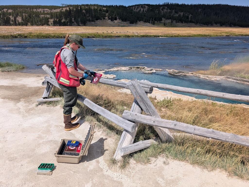 Woman in red vest fastens warning sign to wooden barrier. River, meadow, and forested hill in background.