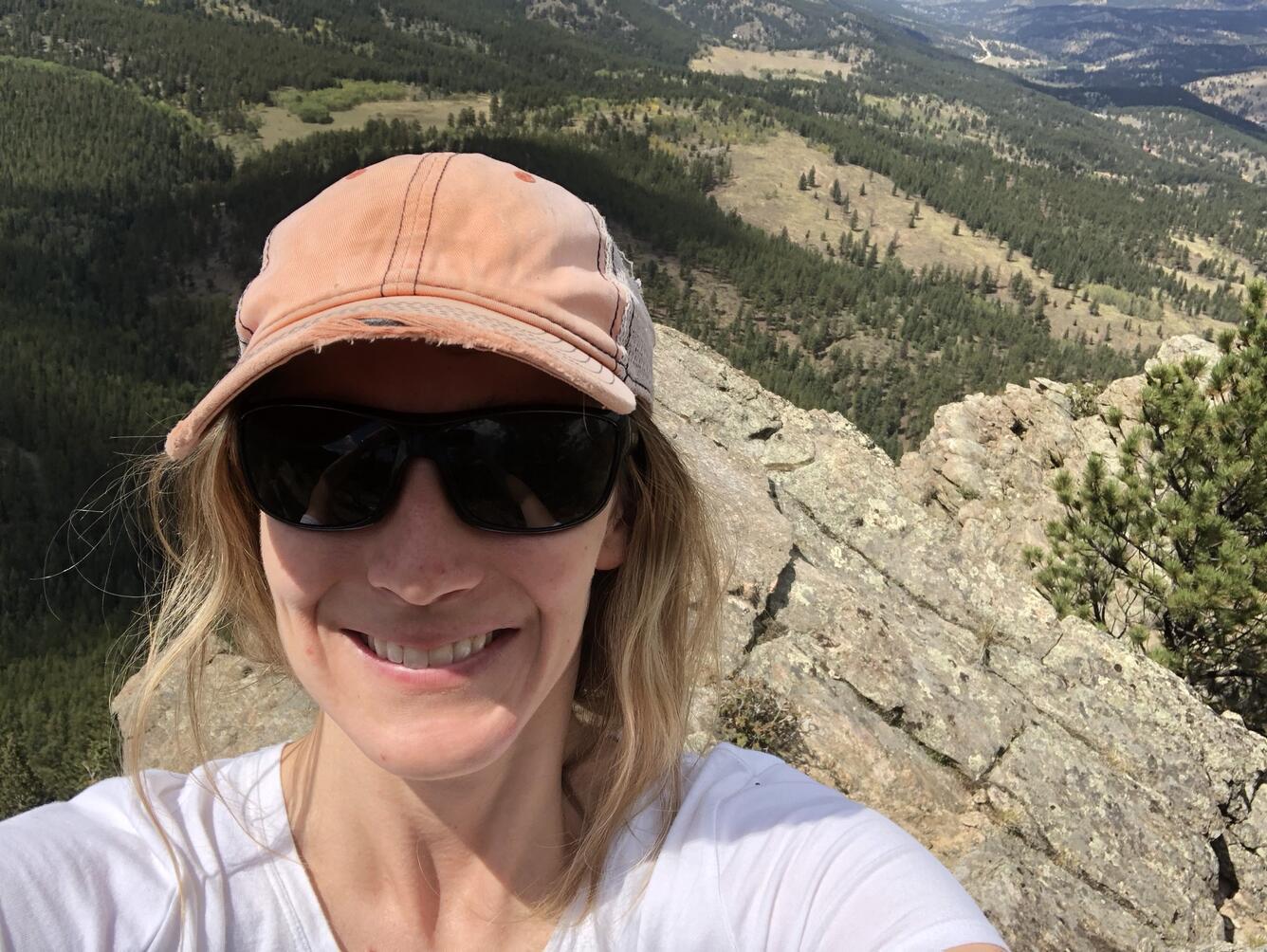 A woman with blonde hair wearing sunglasses and a baseball cap smiles for her picture while on a rocky mountain ridge.