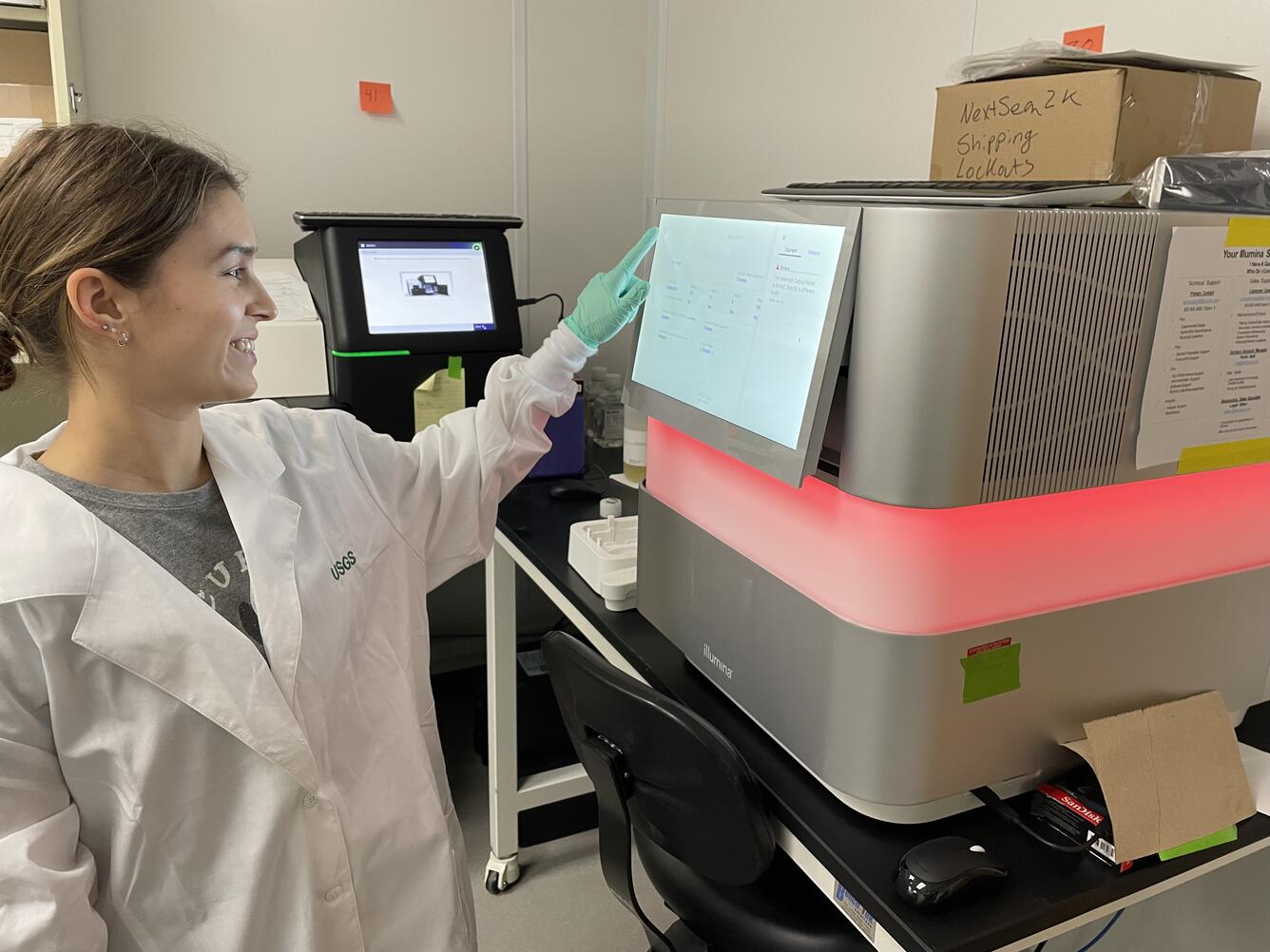 The photograph shows a USGS molecular biologist reviewing the details of a completed high throughput sequencing run on a NextSeq 2000 instrument. She is wearing a lab coat and nitrile gloves. The front of the instrument is visible as well as a MiSeq instrument.