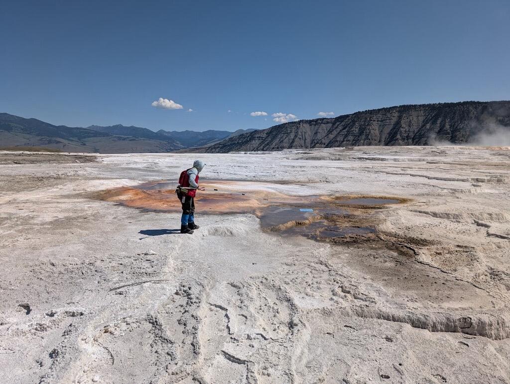 Woman in red vest standing on white terrace in front of reddish hot spring under blue sky. Mountains in the background.