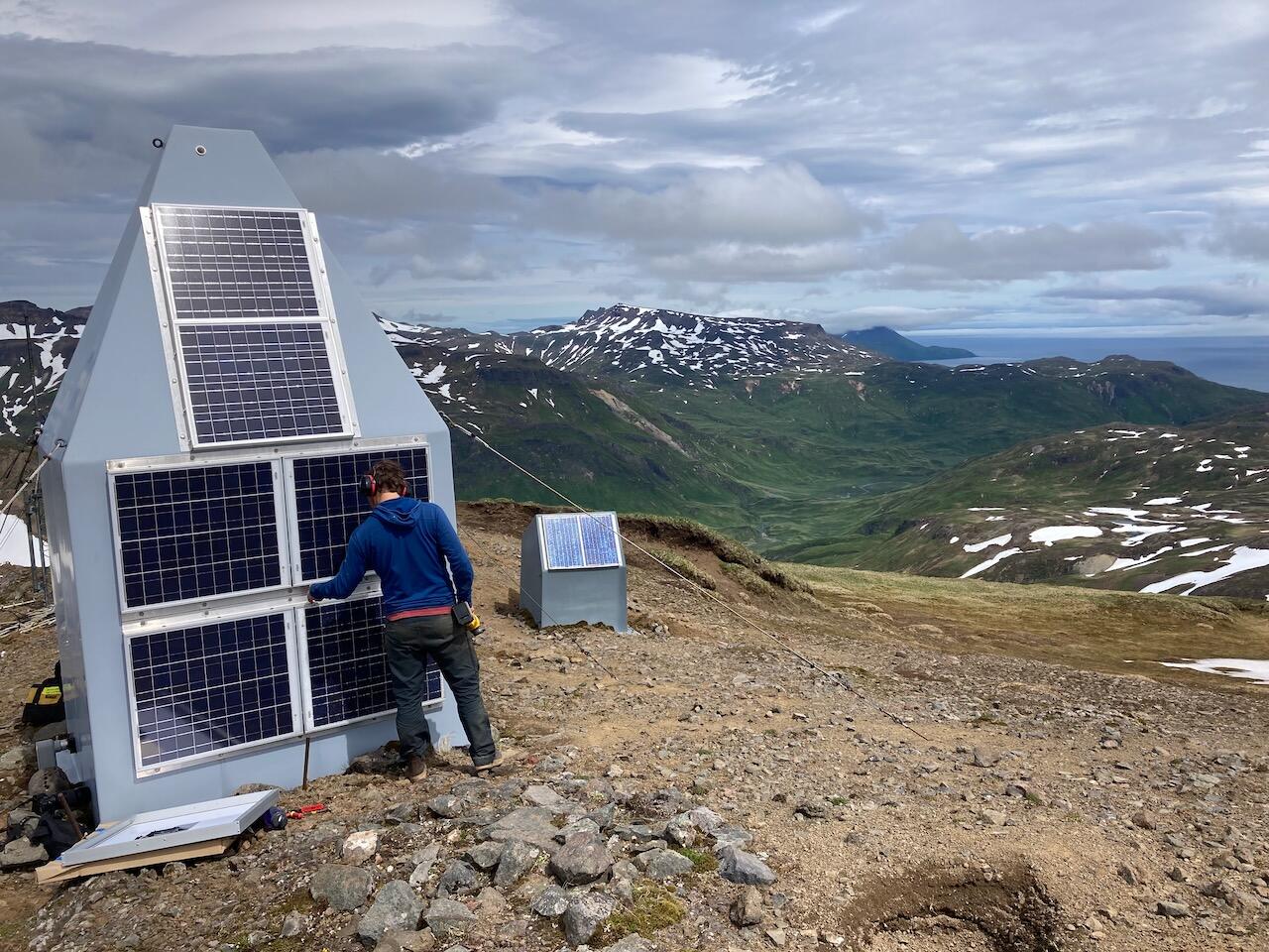 A man stands in front of a small hut installing solar panels, with a valley in the background.