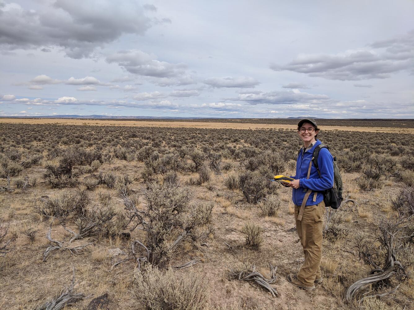 person holding a notebook standing in a field of sagebrush and smiling at the camera