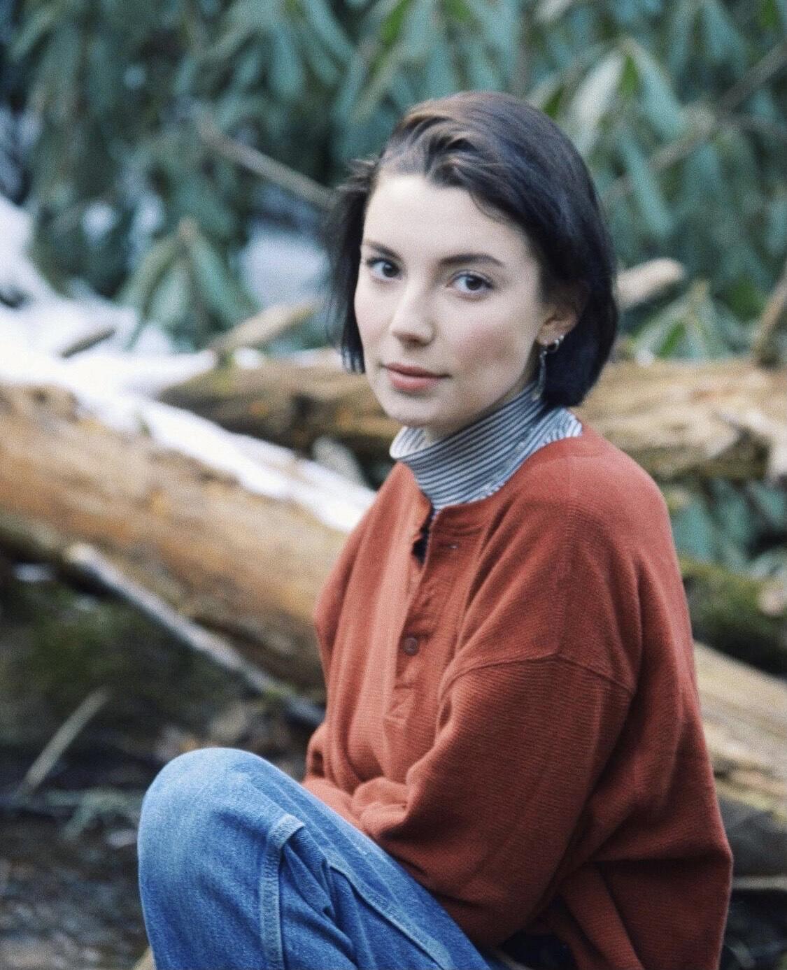 Woman with chin length brown hair wearing red sweater with greenery in background