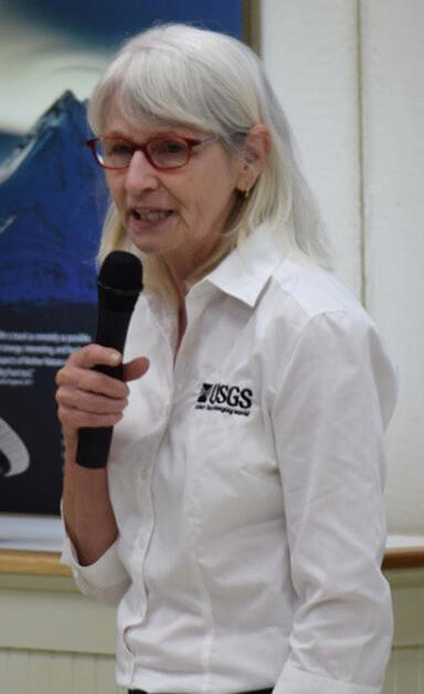 Headshot of a white-haired woman wearing a USGS shirt and red glasses, holding a microphone