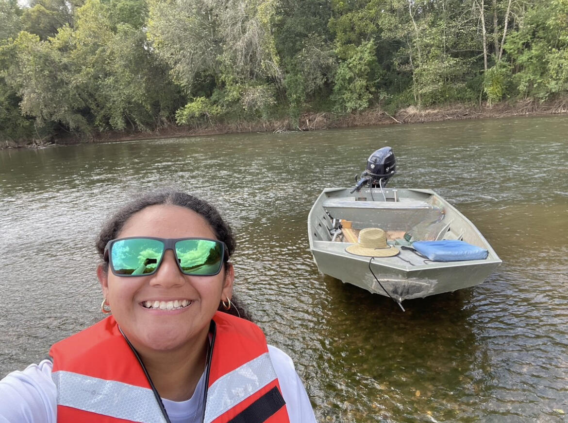 Graduate student with brown hair, wearing green sunglasses and an orange personal floation device, standing near a boat and a