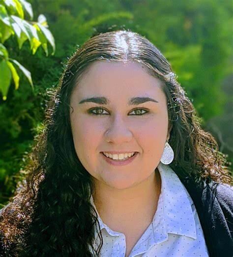 A woman with long dark hair smiles for her picture while wearing a blue button up shirt and a dark blouse. 