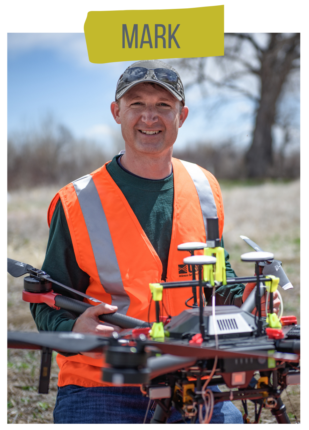 A photo shows USGS scientist Mark Bauer smiling at the camera. Mark is kneeling and holding the USGS drone with both hands. Mark wears a ballcap and an orange vest over his green USGS T-shirt. Behind Mark is a field of brown grass and a leaf-less tree sits in the background.
