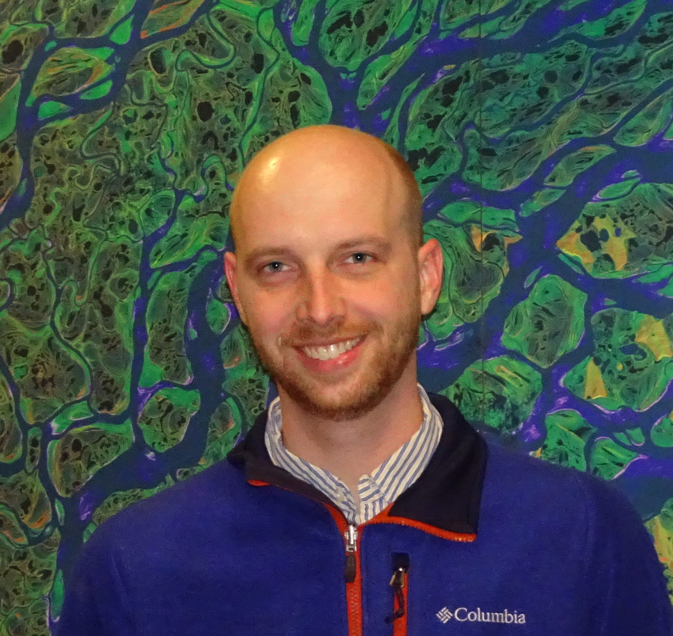 Headshot of a man with a shaved head and a blue fleece, standing in front of a Landsat image