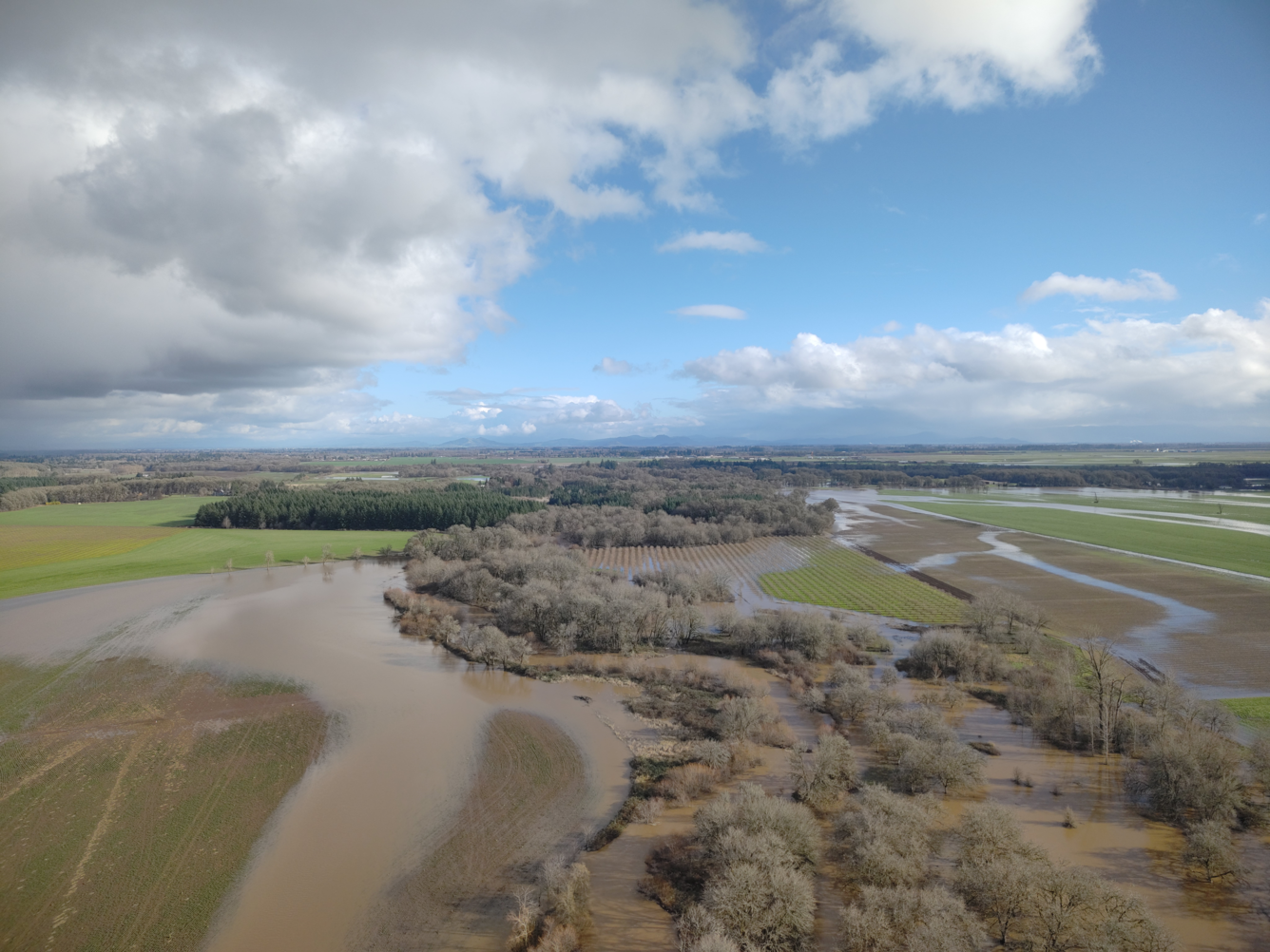 Sediment filled brown meandering river floods into nearby agricultural land. Partly cloudy day.