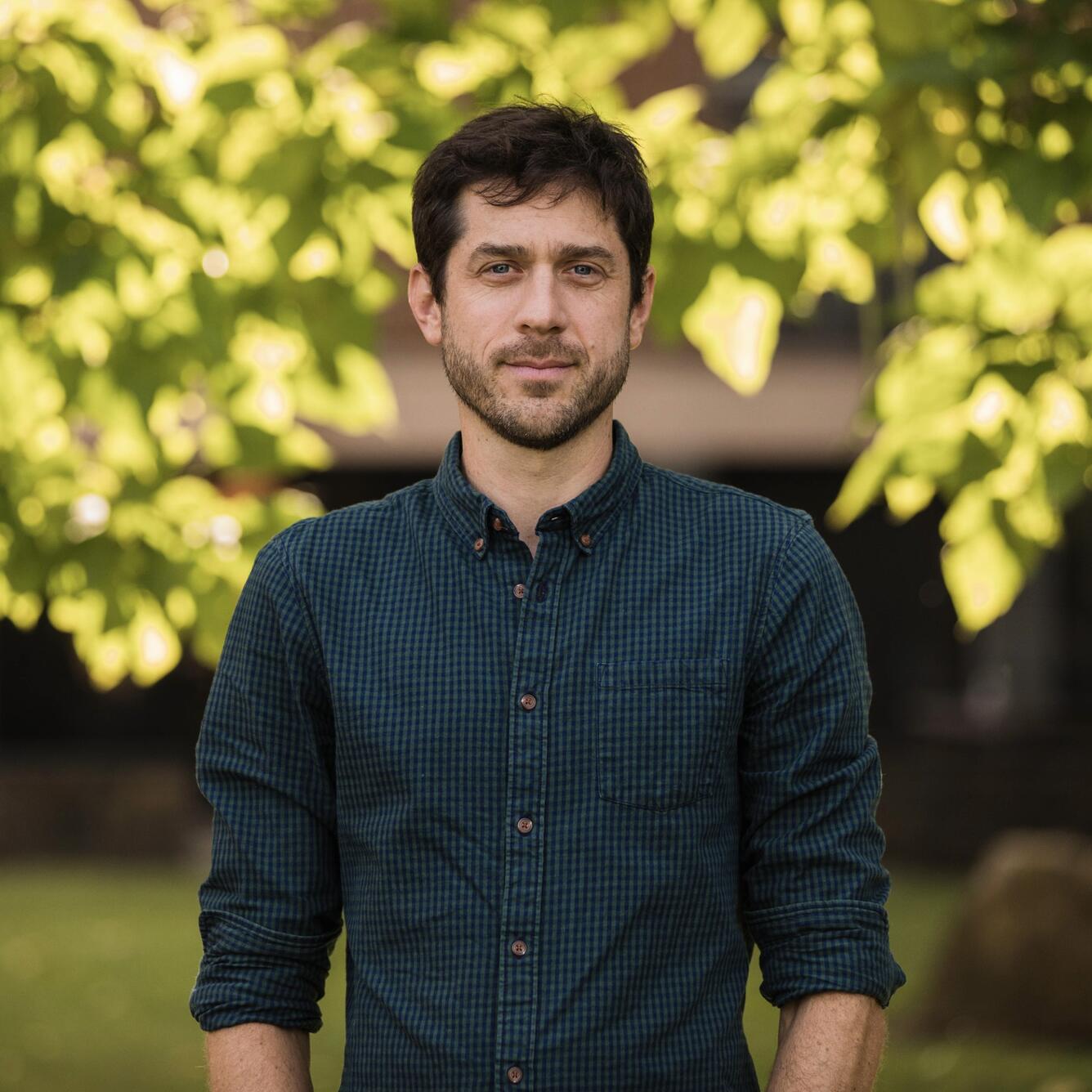 A man with dark hair and wearing a dark green button up shirt stands for his picture.