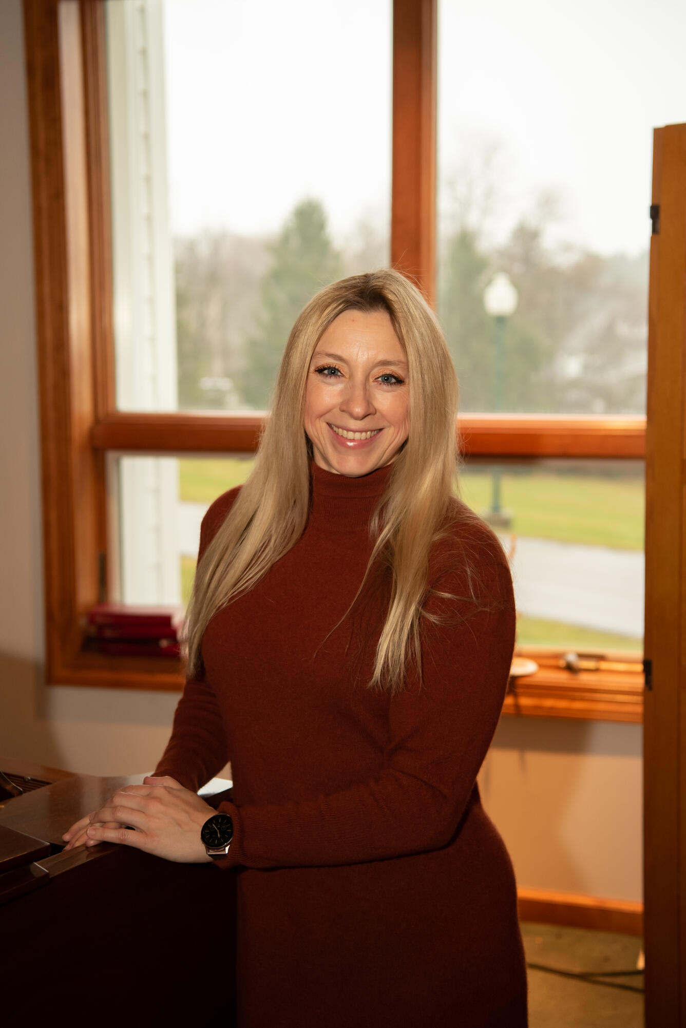 Image of Megan Rogers, smiling, wearing a red sweater and standing in front of a window.