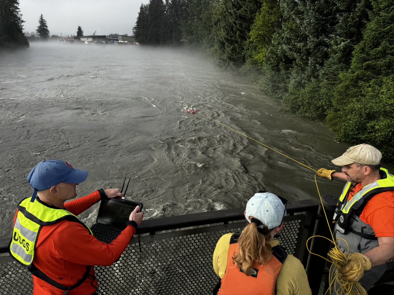 Three scientists stand on a bridge above a flooded river holding a rope attached to scientific equipment in the water below. 