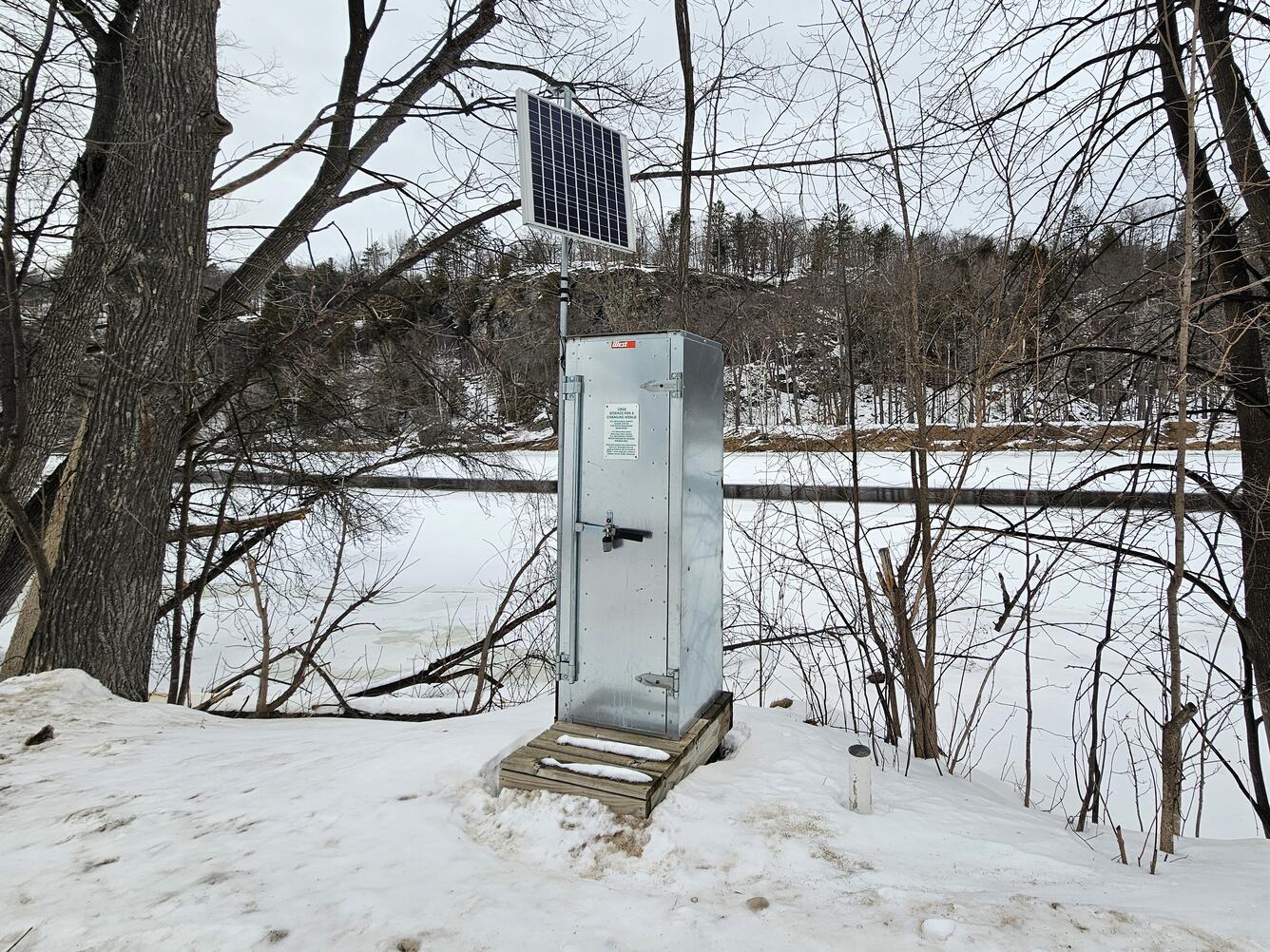A gray metal streamgage housing with an attached solar panel sits on the shore of a snow-covered frozen river.
