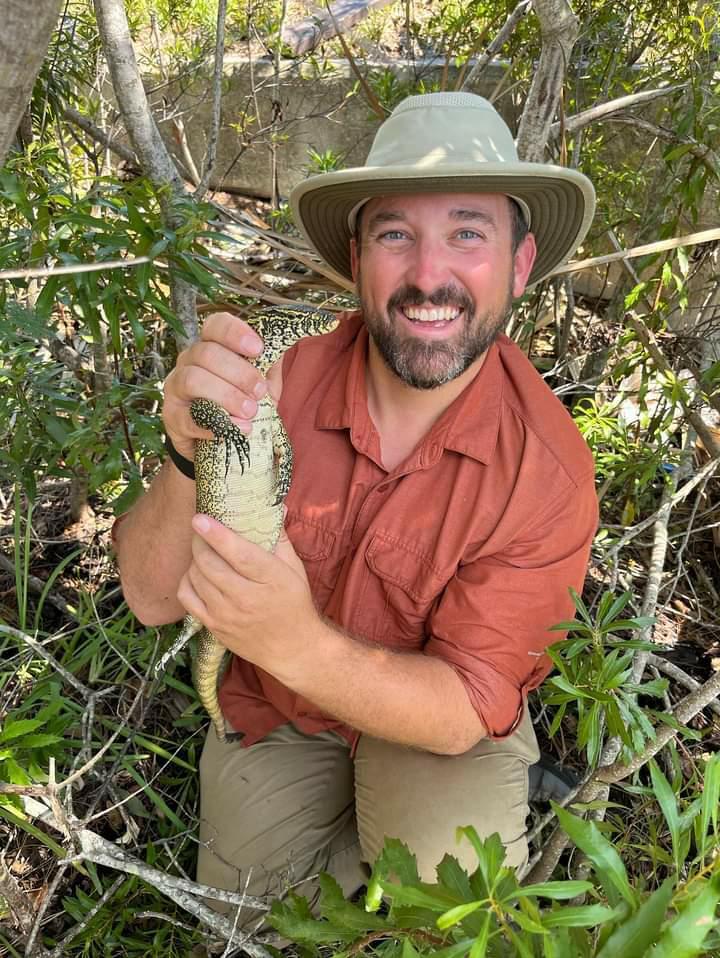 a man in a salmon-colored shirt and a tan hat holds a large black and white lizard, shrubs in the background