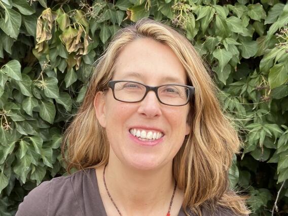 Headshot photo of a smiling woman with shoulder length blond hair and glasses, with vegetation in background