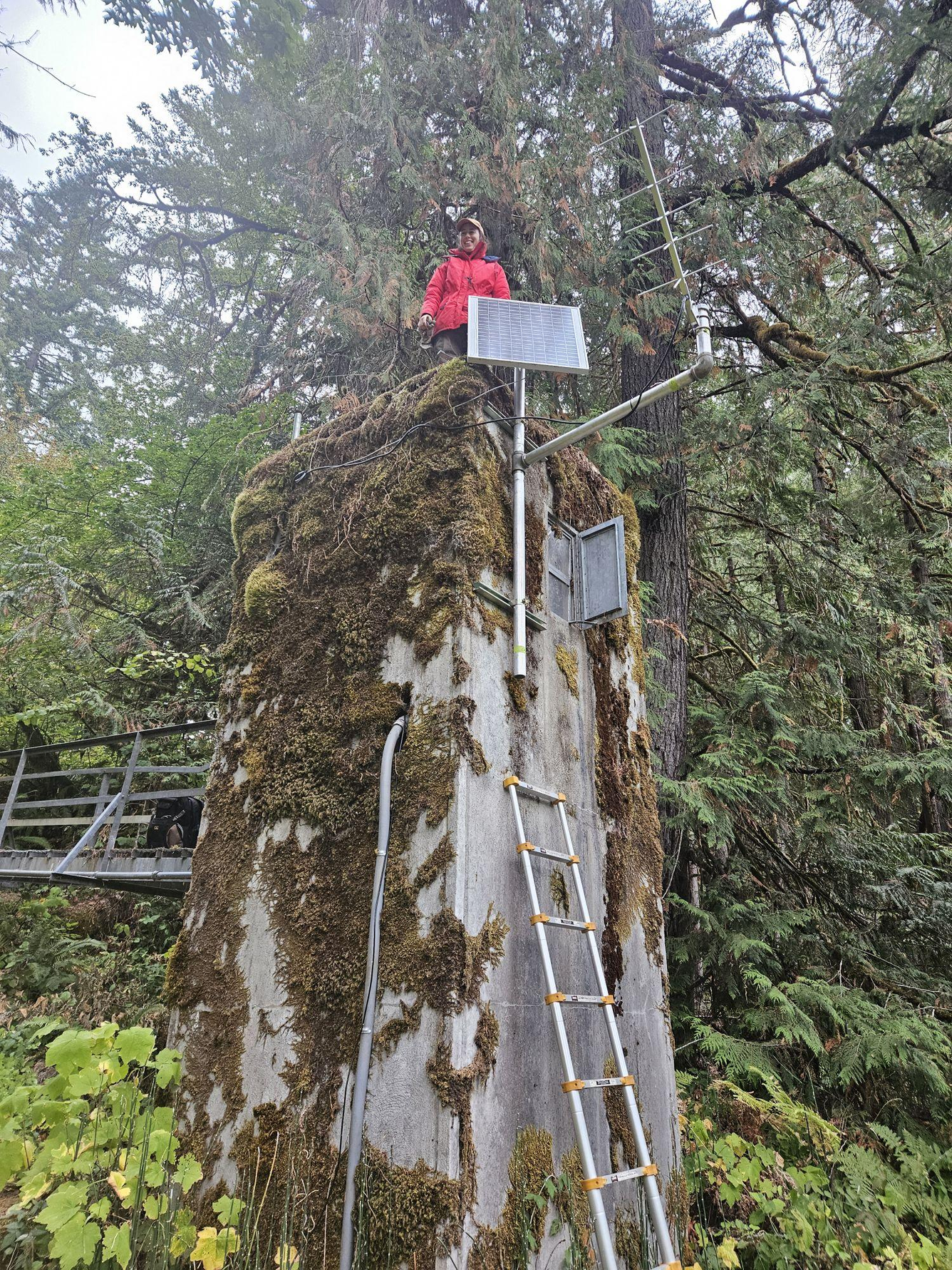 A lady hydro tech in a red coat smiles from on top a mossy gage house.