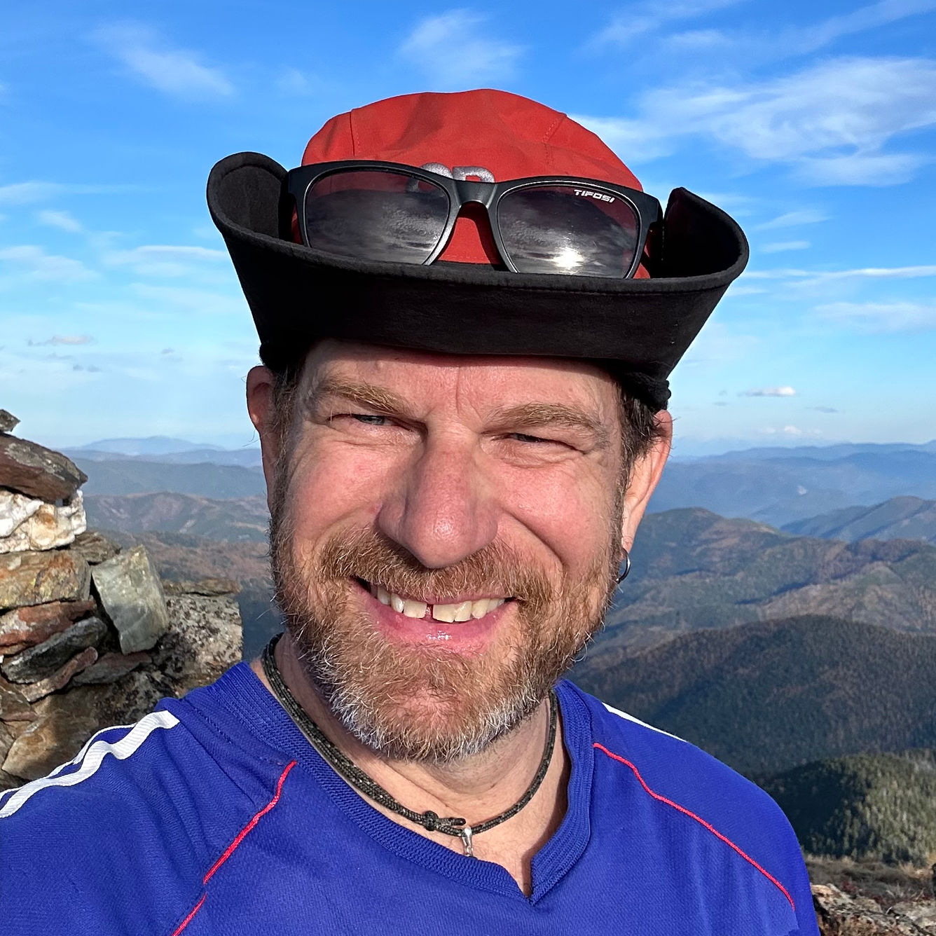 Man wearing red hat with sunglasses on top of hat, smiling with mountains in background