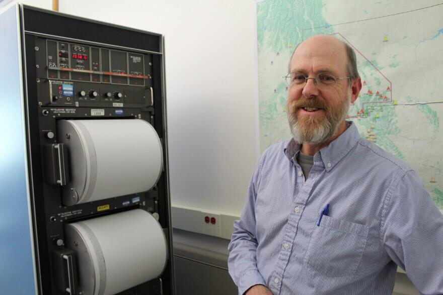 A man stands in front of a machine holding two drums covered in paper that records seismic data