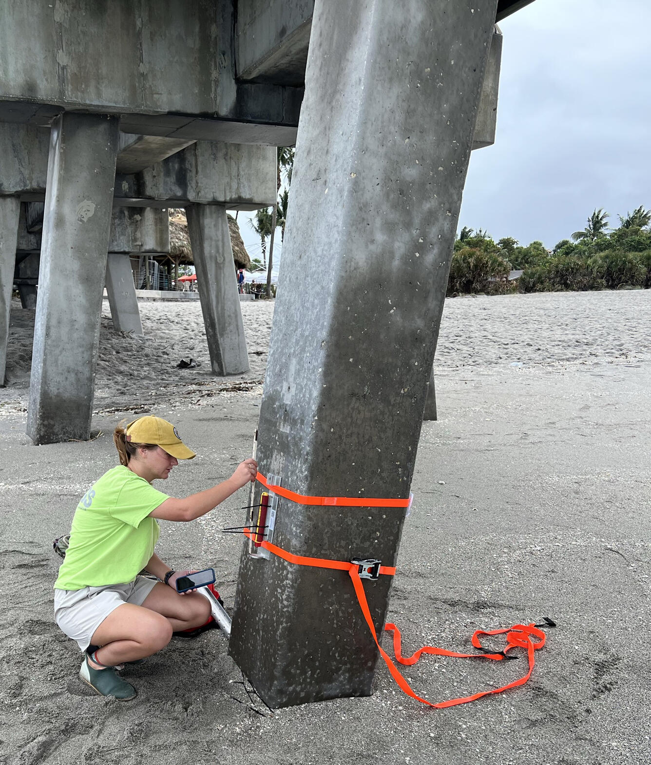 Photo shows a USGS expert on a beach attaching a wave sensor to a concrete pillar under a bridge in Florida. 