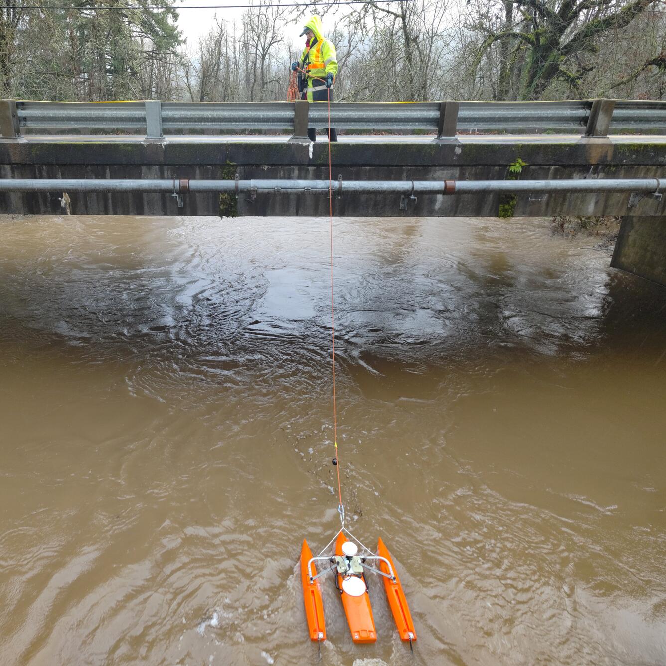 Female in safety gear pulls measurement equipment across the river from a bridge